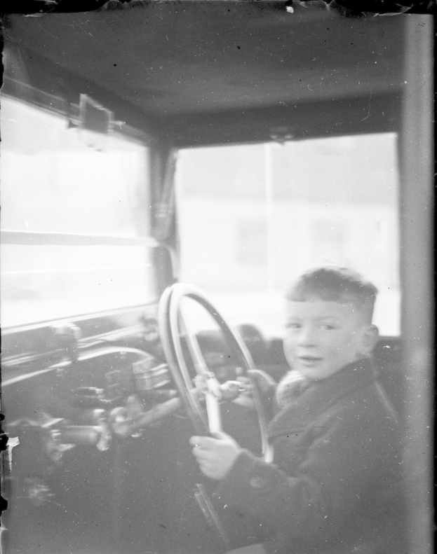 Ein junger Junge in Schwarz-Weiß sitzt auf dem Fahrersitz eines Autos und hält das Lenkrad mit konzentrierter Miene fest.