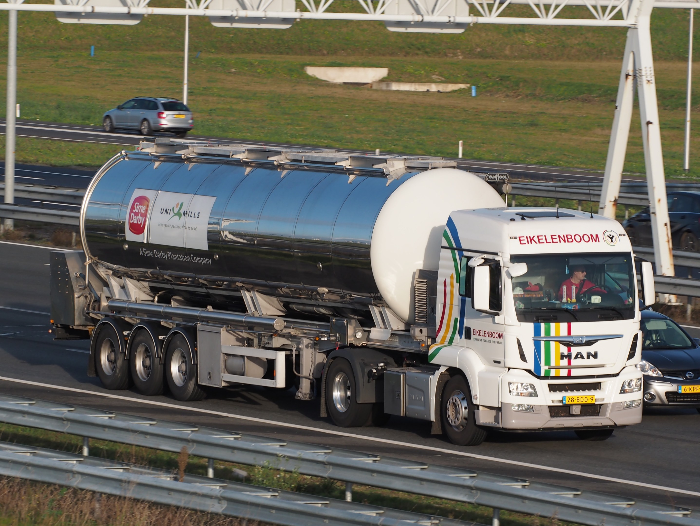 Ein großer Tanklastzug fährt auf einer Autobahn neben einem Auto, mit Menschen im Truck und Gras, Stangen und anderen Gegenständen im Hintergrund.