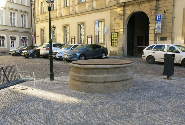 Ein Stadtplatz mit einer zentralen Bank, umgeben von Gebäuden mit Fenstern, Laternenmasten und Schildern sowie einem Springbrunnen in der Mitte.