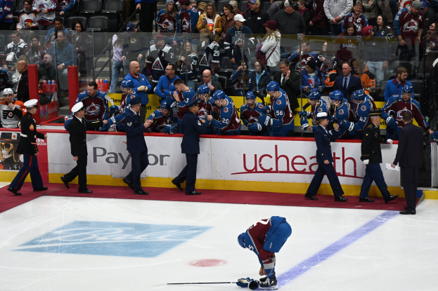 Ein Eishockeyspieler auf dem Eis umgeben von Teamkollegen und Gegnern, mit Zuschauern und Arena-Beschilderung im Hintergrund.