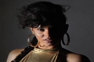 Eine Frau in einem goldenen Kleid und großen Creolen steht selbstbewusst vor einer Wand, ihr glatter Bob-Haarschnitt und das natürliche Make-up unterstreichen ihren entschlossenen Gesichtsausdruck, während sie direkt in die Kamera blickt.