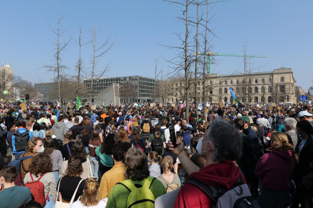 Große Menge von Menschen mit Schildern und Töten vor einem Gebäude mit Bäumen und einem klaren Himmel, was auf eine Klimawandel-Demonstration hinweist.