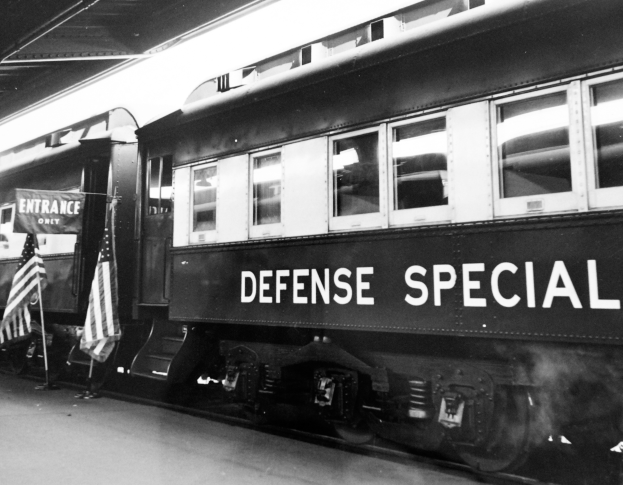 Ein Schwarz-Weiß-Foto eines Zuges mit der Aufschrift 'Defense Special', der auf Gleisen geparkt ist, mit zwei Fahnen links und einer Decke darüber.