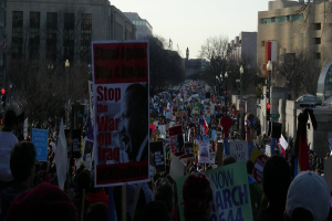 Menschen protestieren auf einer Straße und halten Plakate hoch, mit Gebäuden, Bäumen, Laternenpfählen und dem Himmel im Hintergrund.