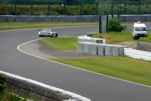 Ein Rennauto fährt auf einer Rennstrecke mit einem Polizeiwagen im Hintergrund, umgeben von Gras, Pflanzen, Bäumen und Zaun, mit einem Fahrzeug auf der rechten Seite und Text unten.