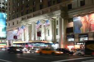 Eine nächtliche Stadtstraße mit fahrenden Fahrzeugen, ein Gebäude mit Fenstern, Säulen und Fahnenstangen, beleuchtete Schilder mit Text und einem Frauenbild, sowie umgebende Lichter.