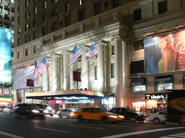 Eine nächtliche Stadtstraße mit fahrenden Fahrzeugen, ein Gebäude mit Fenstern, Säulen und Fahnenstangen, beleuchtete Schilder mit Text und einem Frauenbild, sowie umgebende Lichter.
