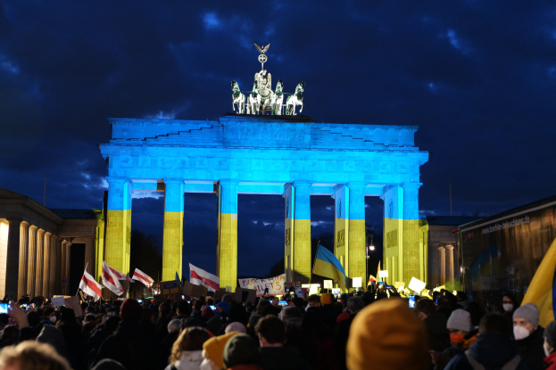 Menschenmenge mit Fahnen und Plakaten vor dem Brandenburger Tor in Berlin, mit einer Fahne auf der rechten Seite.