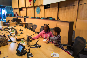 Zwei Kinder sitzen an einem langen Tisch mit Mikrofonen, Flaschen, Taschen und Papieren und diskutieren bei einer Schulratssitzung, umgeben von Menschen und Flaggen im Hintergrund.