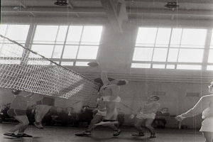Eine Gruppe von Menschen, die Volleyball in einer Turnhalle mit einem Netz in der Mitte spielt und Zuschauer auf Tribünen im Hintergrund sitzen, dargestellt in Schwarz-Weiß.