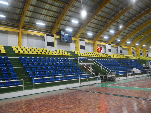 Großer Hallensportplatz mit Holzboden, blauen und gelben Sitzplätzen, Geländern, Treppen zum Sitzbereich, wandmontierten Bildschirmen, Deckenleuchten und einigen Menschen.