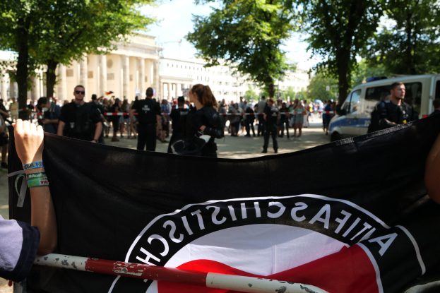 Gruppe von Menschen, die auf dem Boden stehen und eine deutsche Flagge vor einer Menge halten, mit einem Metallstab, Fahrzeugen, einem Zaun, Bäumen, Gebäuden mit Fenstern und einem bewölktem Himmel im Hintergrund.