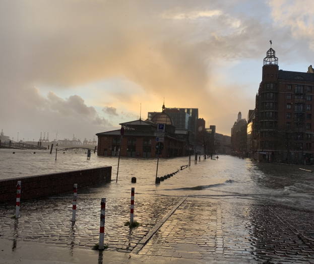 Uberflutete Straße in Hamburg, Deutschland mit Wasser bedeckt, Pfölen, Schilder, Gebäude und eine Brücke unter einem bewölktem Himmel.