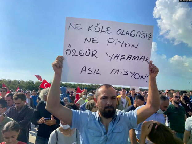 Ein Mann hält ein Schild mit der Aufschrift "Aserbaidschanische Demonstranten demonstrieren in Ankara für den Rücktritt von Präsident Ilham Aliyev" vor einer großen Menge mit Fahnen und Handys, unter einem blauen und weißen Himmel mit Bäumen im Hintergrund.