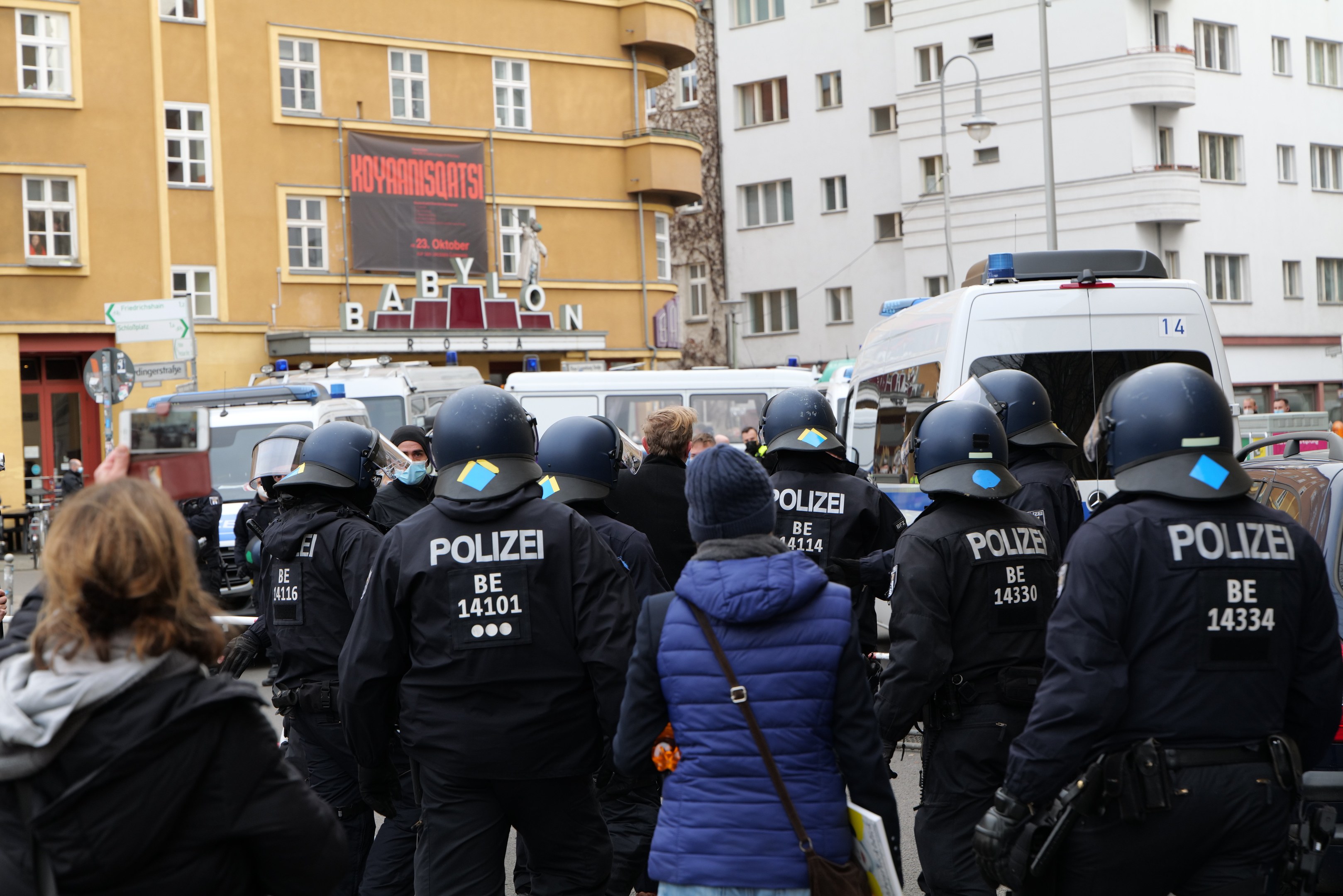 Eine Gruppe von Polizisten in Uniform steht vor einer Menge von Menschen mit Helmen und Jacken während einer Demonstration in Berlin, Deutschland, mit Fahrzeugen, Gebäuden, Laternenmasten und einem Banner im Hintergrund.