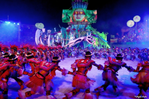 Eine Gruppe von Menschen in bunten Kostümen tanzt auf einer Bühne vor einem sitzenden Publikum, beleuchtet von Lichtern und umgeben von Polen und Ballons, mit einem Bildschirm, der eine Person und andere Gegenstände zeigt, im Hintergrund, während des Rio-Karnevals 2014.
