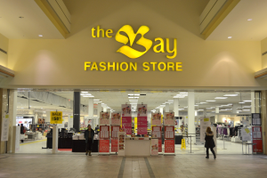 Außenansicht eines Bay Fashion Stores in einem Einkaufszentrum mit Kunden, Wandtexttafeln, Säulen, Deckenleuchten und Hintergrundauslagen.