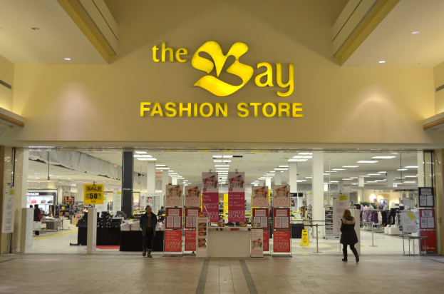 Außenansicht eines Bay Fashion Stores in einem Einkaufszentrum mit Kunden, Wandtexttafeln, Säulen, Deckenleuchten und Hintergrundauslagen.