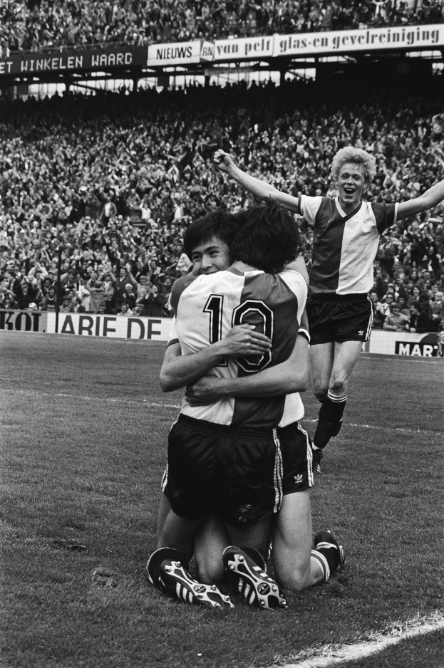 Drei Fussballspieler umarmen sich auf dem Boden und feiern ein Tor vor einer jubelnden Menge in einer schwarz-weißen Stadion-Szene.