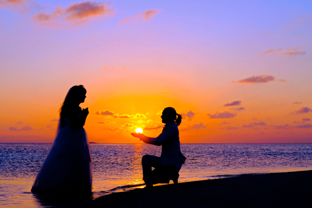Braut und Bräutigam am Strand bei Sonnenuntergang, Braut in einem weißen Kleid, Bräutigam kniend mit einem Gegenstand in der Hand, leuchtendes Orange, Pink und Purpur am Himmel, tiefes Blau im Wasser.