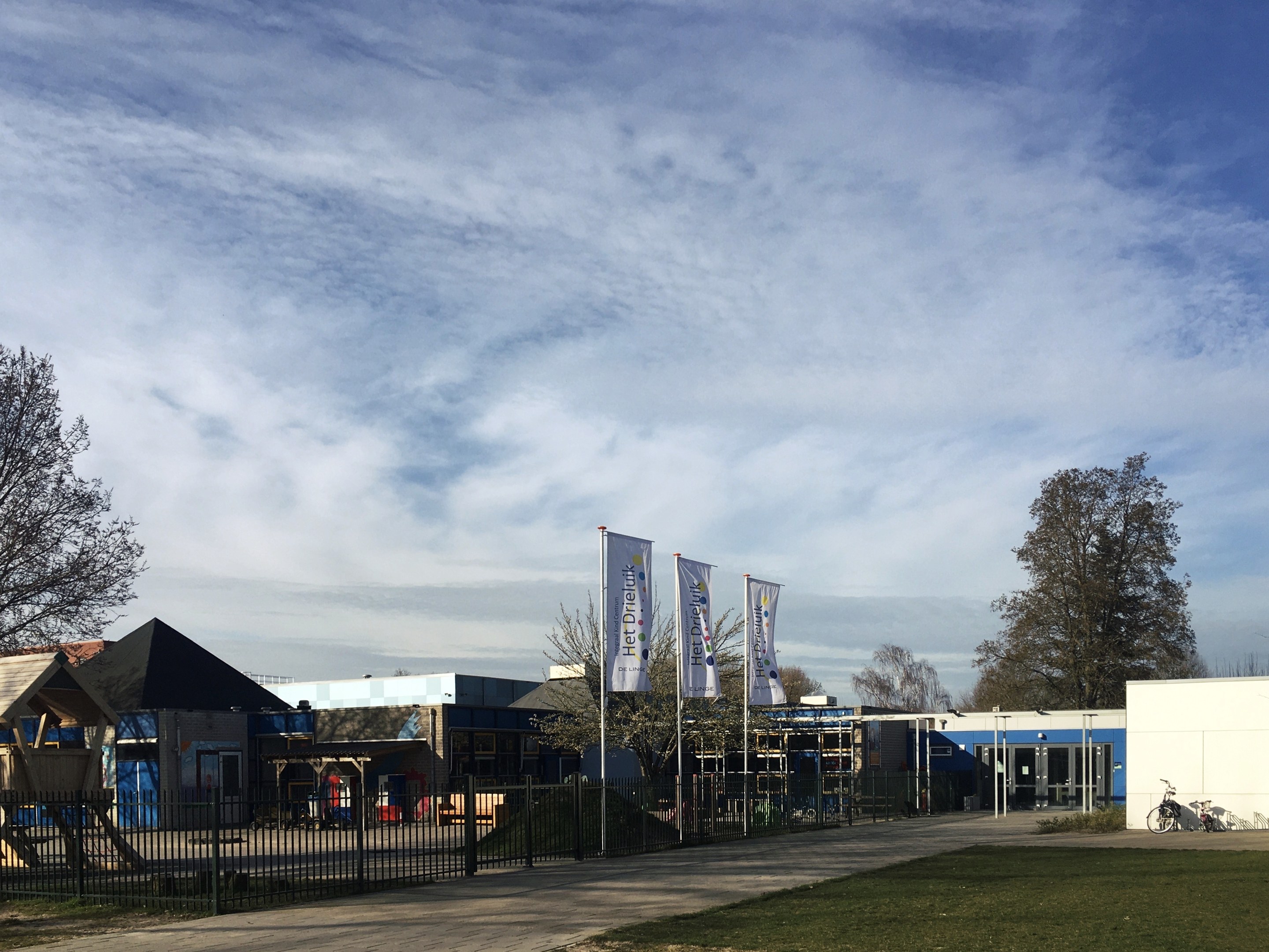 Großes Backsteinschulgebäude mit einem Rasenplatz, der von einem Metallzaun umgeben ist, mit einem Weg, Bäumen, Bannern, einem geparkten Fahrrad, nahen Häusern und einem bewölkten Himmel.