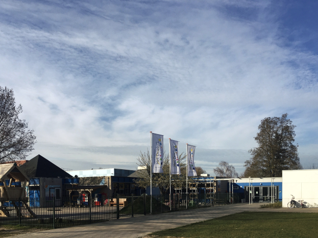 Großes Backsteinschulgebäude mit einem Rasenplatz, der von einem Metallzaun umgeben ist, mit einem Weg, Bäumen, Bannern, einem geparkten Fahrrad, nahen Häusern und einem bewölkten Himmel.