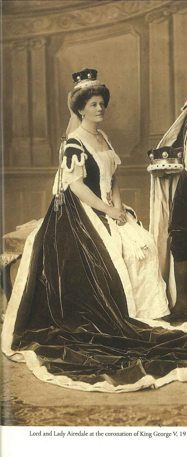 Lady Airedale, in einem schwarzen und wei├čen Kleid, steht vor einer Wand mit einem Objekt rechts davon und Text unten im Bild.
