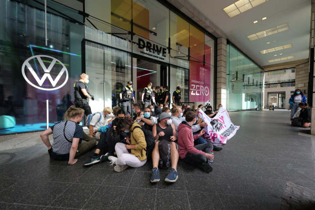 Eine Gruppe von Menschen sitzt vor einer Volkswagen-Vertretung mit Glaswänden auf dem Boden und hält eine Schilder mit Text, unter Deckenbeleuchtung, scheinbar protestierend.