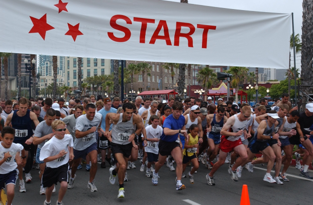 Gruppe von Läufern bei einem Marathon mit einem Verkehrskegel im Vordergrund und einer Fahne im Hintergrund, umgeben von Bäumen, Laternenmasten, Gebäuden und einem klaren blauen Himmel.