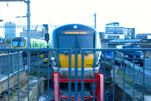 Zwei Züge auf dem Gleis mit einem Zaun im Vordergrund, Pfosten auf dem Land, einer mit einer Signalleuchte, ein Auto auf der Straße rechts, Gebäude im Hintergrund und der Himmel darüber.