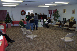 Ein Raum mit mehreren Stühlen, Tischen und Personen, einige stehen, einige sitzen. Auf den Tischen stehen Tassen und Gefäße. Im Hintergrund eine Wand mit einem Strauß, Vorhängen, Tür, Fotorahmen und Fenstern. Ein Weihnachtsbaum und ein Piano auf der rechten Seite. Leuchtstoffröhren an der Decke.