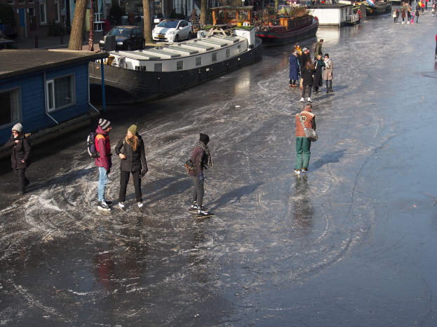 Eine Gruppe von Menschen, die auf einem zugefrorenen Kanal in Amsterdam Eislaufen, umgeben von Gebäuden, Bäumen, Pfählen, Fahrzeugen und anderen Gegenständen, mit dem Himmel im Hintergrund und dem Wasser, das in der Sonne glitzert.
