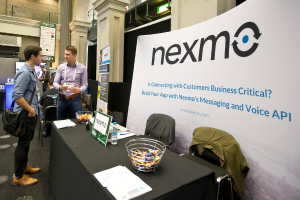 Zwei Personen stehen links, eine trägt eine Tasche und die andere trägt einen Ausweis, mit einem Tisch in der Mitte mit Papieren, Körben, einer Karte und einer Flasche; im Hintergrund eine Tafel mit Text und Säulen und eine Gruppe von Personen links.