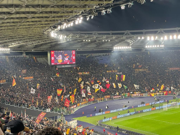 Eine große Menge in einem Stadion schwenkt Fahnen, mit Bannern, einem Bildschirm und beleuchteter Decke, was auf Spannung für ein Fußballspiel hindeutet.