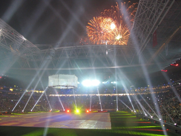 Olympiastadion in London bei Nacht, erhellt von hellen Lichtern, umgeben von einer großen Menge, mit grünem Gras und Feuerwerk, das den Himmel erleuchtet.