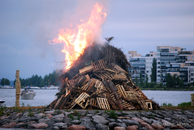 Holzobjekte brennen mit Feuer, Steine unten, Schiff auf Wasser in der Mitte, mit Geb√§uden, B√§umen und Himmel im Hintergrund.