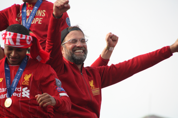 Liverpool-Trainer Jürgen Klopp feiert mit seinen Spielern den Gewinn der UEFA Champions League, während zwei medaillentragende Personen in der Nähe stehen und der Himmel im Hintergrund zu sehen ist.