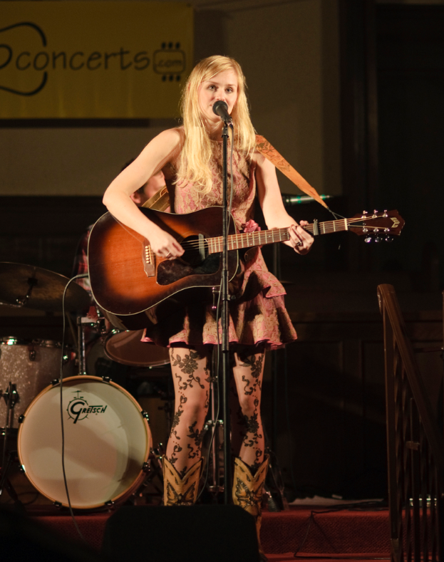 Eine Frau, die auf der BÃ¼hne eine Gitarre spielt und singt, mit einer Band im Hintergrund und einem Bildschirm an der Wand hinter ihr.