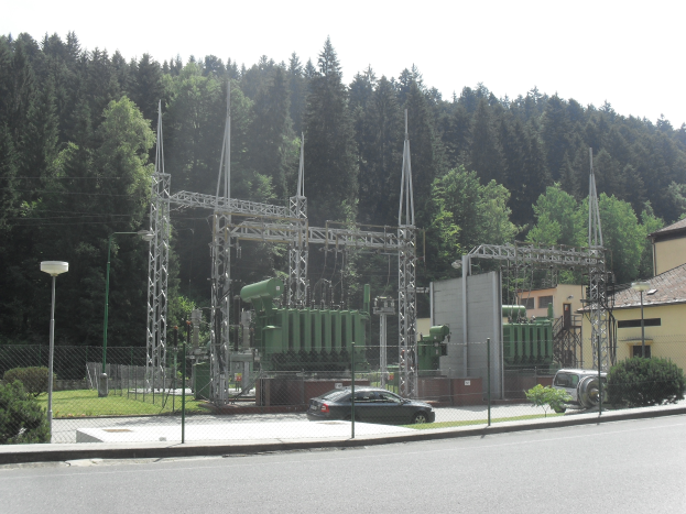 Ein elektrisches Umspannwerk am Straßenrand, umgeben von einem Zaun, Masten, Laternen, Pflanzen, Gras und Bäumen, mit einem vorbeifahrenden Auto und Gebäuden im Hintergrund unter einem klaren blauen Himmel.