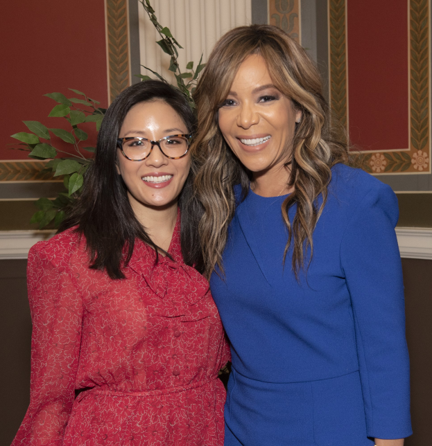 Zwei Frauen, die bei der 2019er Women in Film Pre-Oscar-Luncheon zusammen lächeln, eine in einem blauen Kleid und die andere in einem roten Kleid mit Brille, stehen neben einer Pflanze und einem Vorhang.