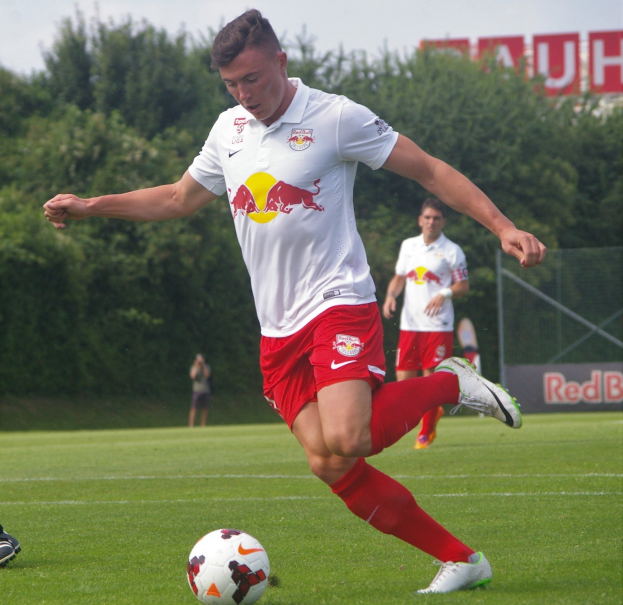 Ein professioneller Fußballspieler in einem roten Trikot kickt einen Ball auf einem grasbewachsenen Feld mit Bäumen und einem klaren blauen Himmel im Hintergrund, in der Nähe einer Tafel mit der Aufschrift "RB Leipzig."