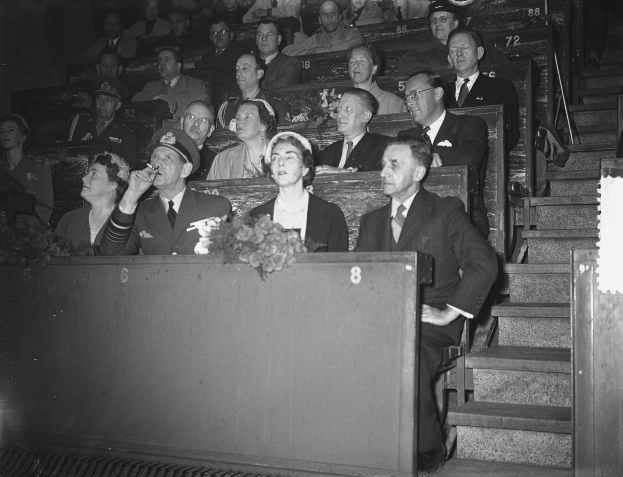 Schwarzes und weißes Foto einer Zuschauermenge in einem Stadion mit einem Podium und einem Blumenstrauß davor, Treppen auf der rechten Seite.