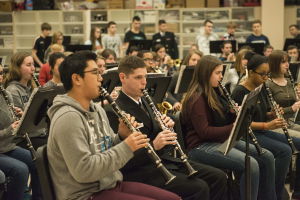 Eine Gruppe von Menschen, die Klarinetten in einem Klassenzimmer spielen, einige sitzen und einige stehen, und Regale mit Kisten im Hintergrund.