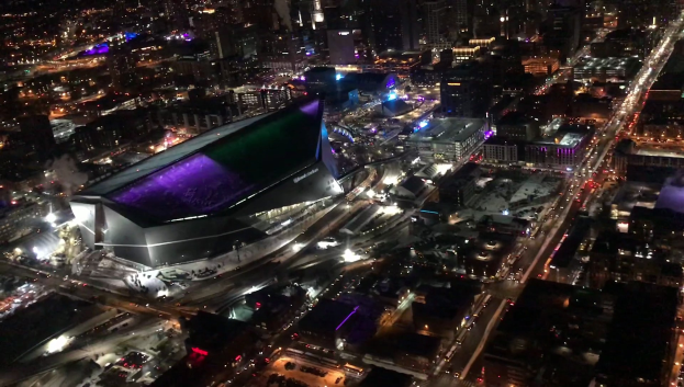 Luftaufnahme einer hell erleuchteten Stadt in der Nacht mit einem großen zentralen Stadion, umgeben von beleuchteten Gebäuden und befahrenen Straßen.