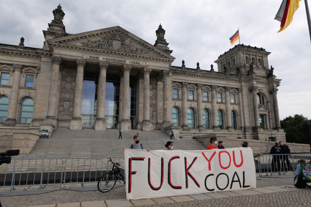 Gruppe von Menschen mit einem "Fuck You Coal"-Schild vor dem Reichstag in Berlin, mit seinen architektonischen Details, einem Fahrrad, Bäumen, einem Fahnenmast und bewölktem Himmel.