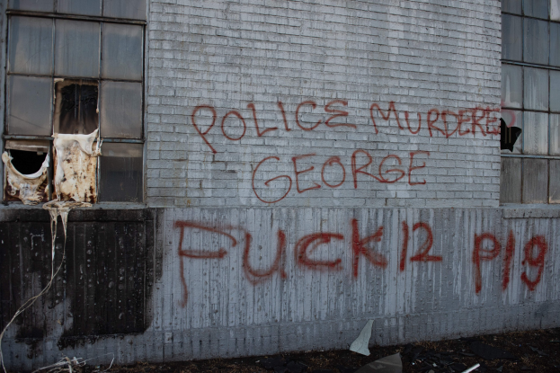 Graffiti an einem Gebäude mit der Aufschrift "Polizei ermordet George Fuck 12 P19" und zerstreuten Gegenständen davor.