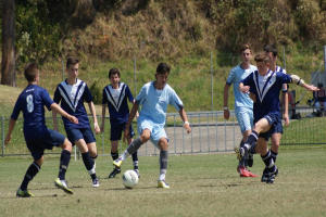 Eine Gruppe von Menschen spielt Fußball mit einem Ball auf dem Boden, während zwei Personen im Hintergrund auf Stühlen sitzen und weiter weg Bäume zu sehen sind.