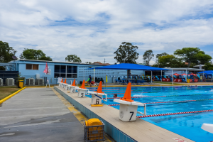 Ein großes Schwimmbad mit schwimmenden Menschen, Bahnteiler, Verkehrskegeln, Stühlen, Sonnenschirmen, einem Gebäude mit Fenstern, einer Flagge, Bäumen und einem bewölkten Himmel im Hintergrund.