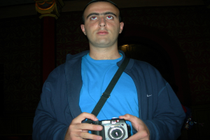 Ein Mann steht mit beiden Händen eine Kamera haltend da und trägt ein Bank-T-Shirt.