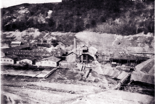 Ein Schwarz-Weiß-Foto eines Kohlebergwerks mit einigen Gebäuden, Masten und Bäumen im Hintergrund und Text am unteren Rand.